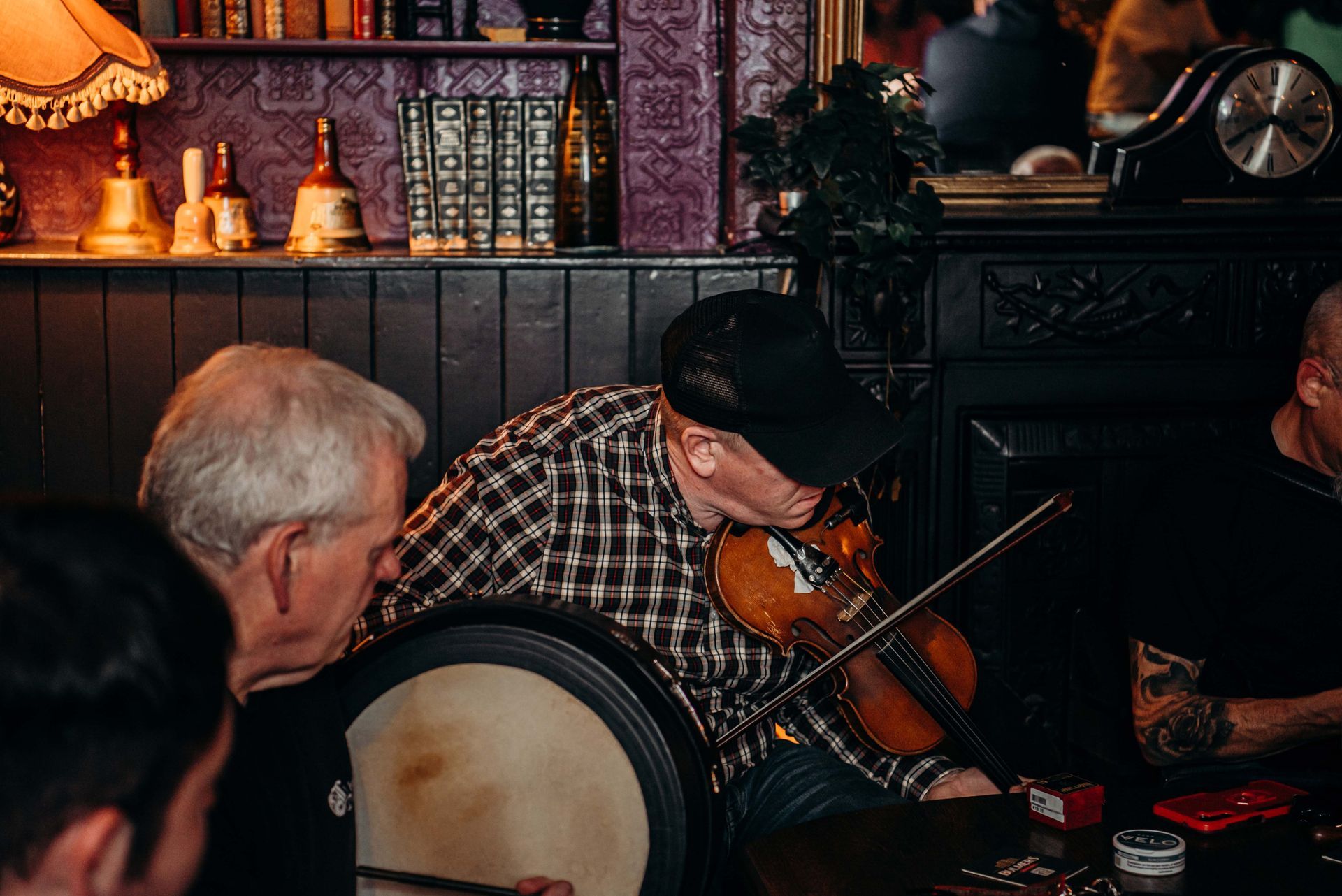 Musicians in pub playing instruments: fiddle, drum. Dark setting, bookshelves, dim lighting.