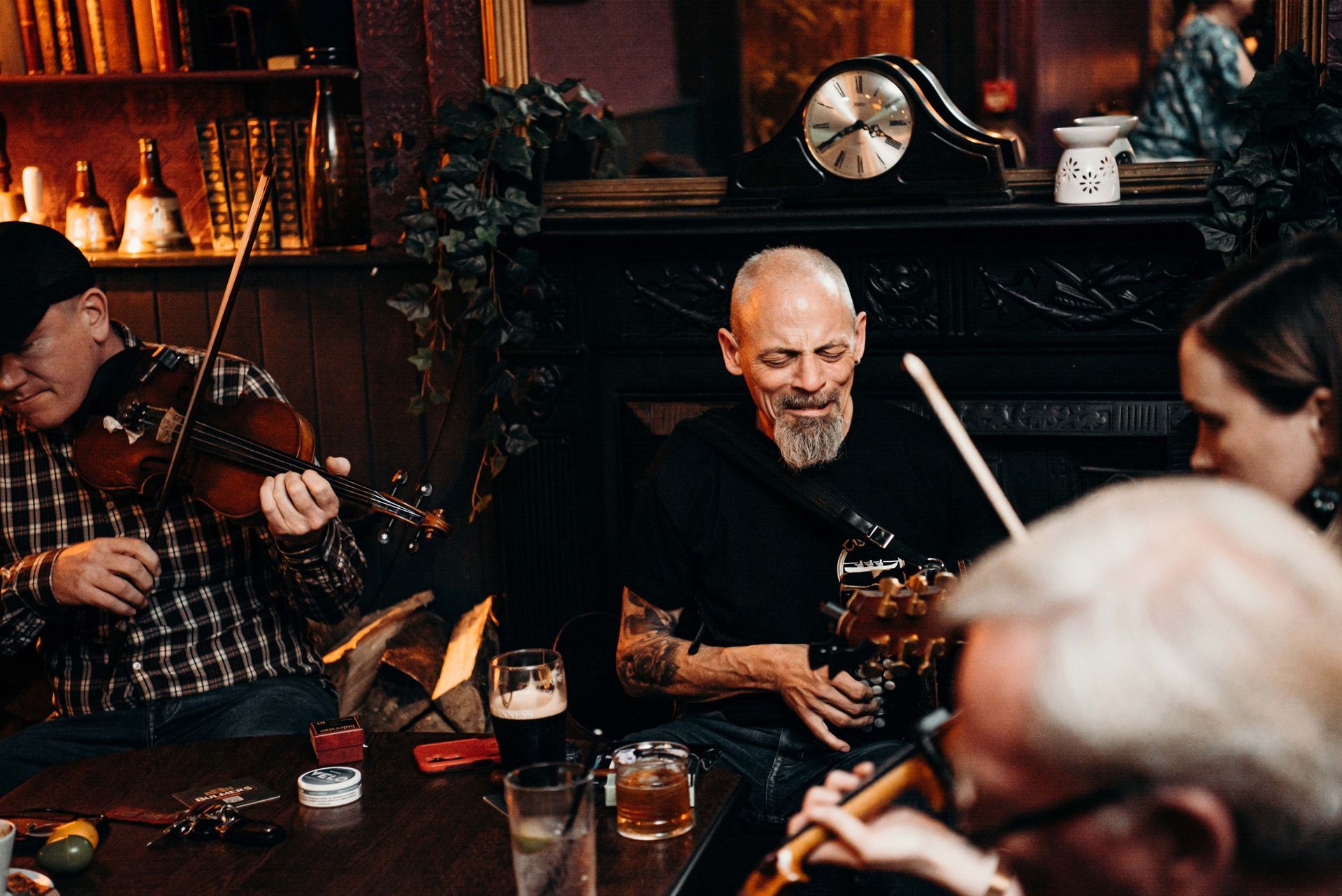 Musicians playing instruments in a dimly lit bar. Man with tattoos sings while others play the fiddle and flute.