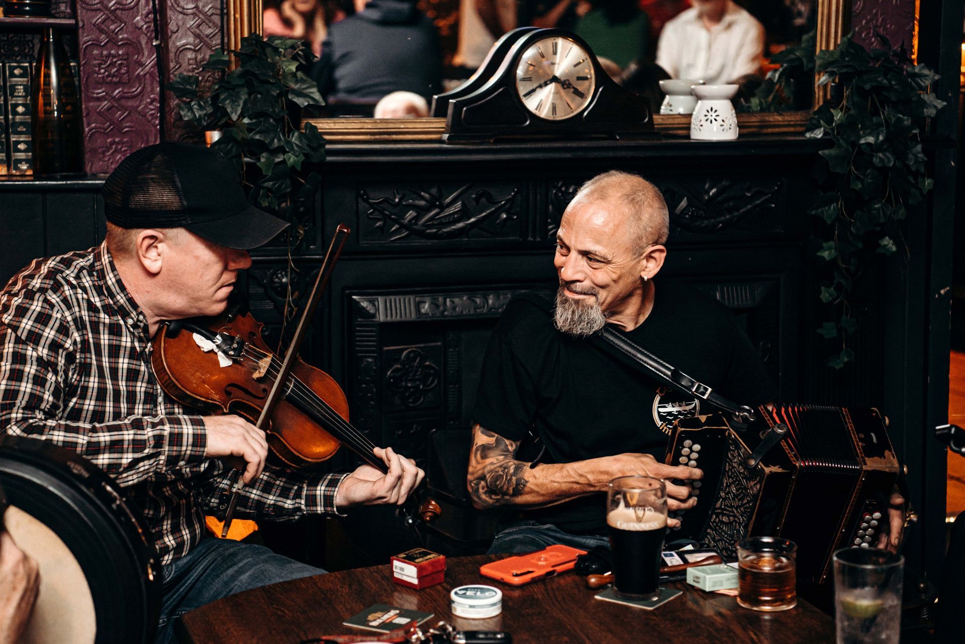 Two musicians in a pub, playing violin and accordion. One with a cap, the other with a beard.