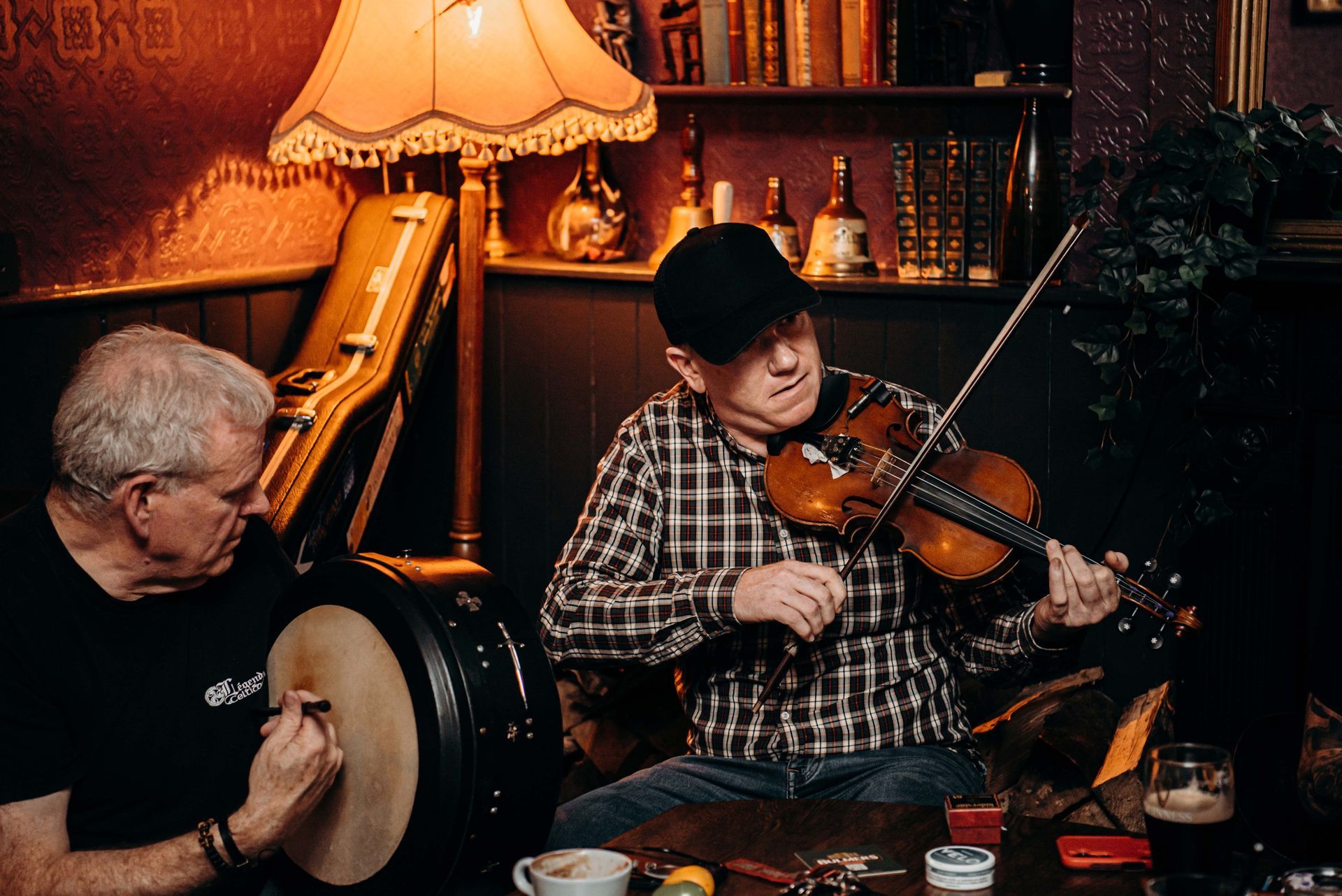 Two musicians playing in a dimly lit pub; one with a fiddle, the other with a bodhrán.
