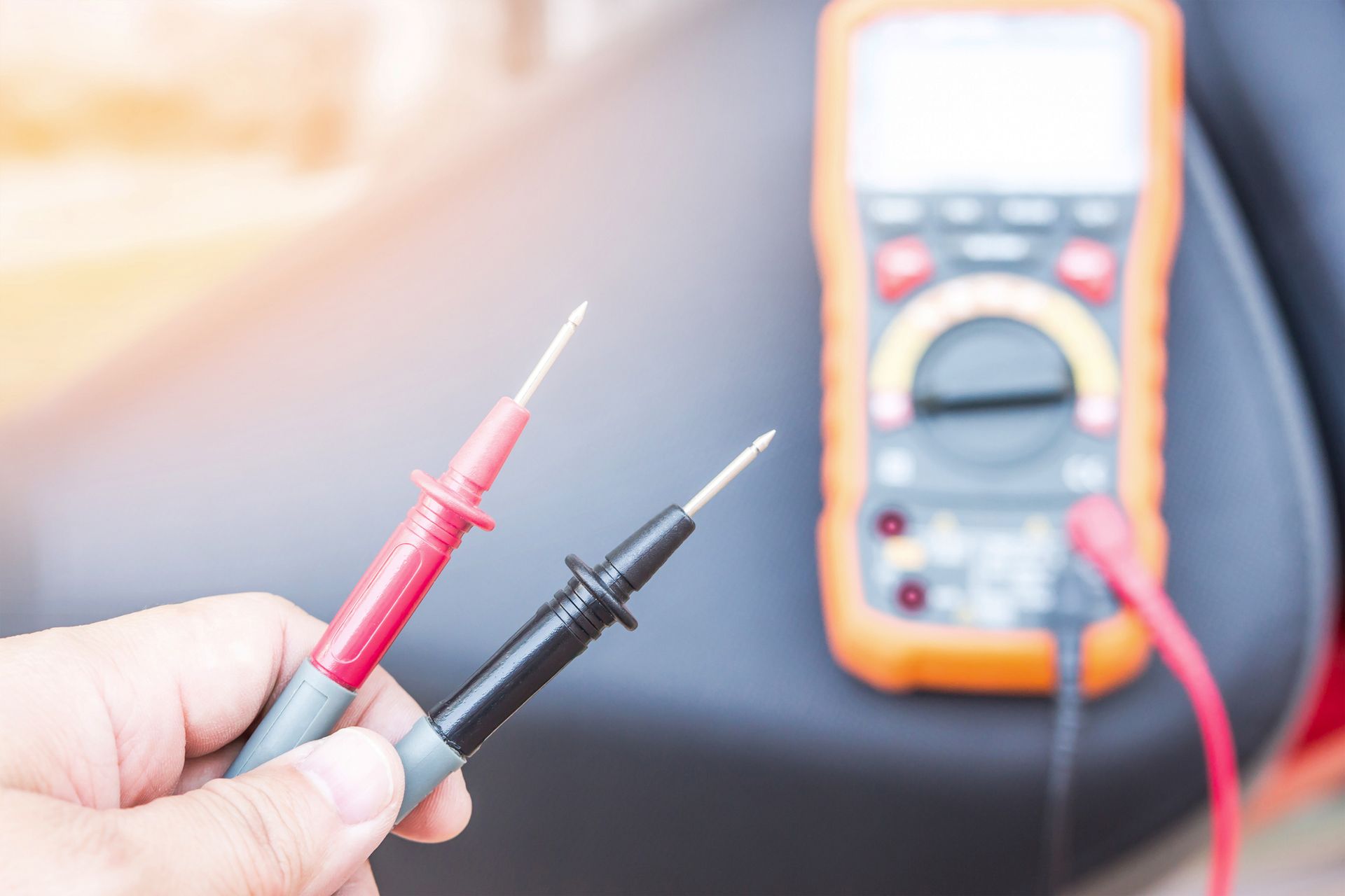 A Person Is Holding Two Test Probes in Their Hand in Front of A Multimeter.