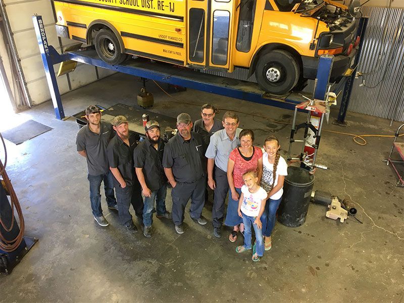 Group of people posing in a garage with a school bus lifted on a hoist. | Mohr's Automotive