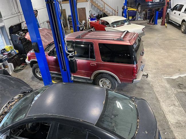Red SUV and black car in a repair shop, other vehicles, a lift, and equipment are visible. | Mohr's Automotive