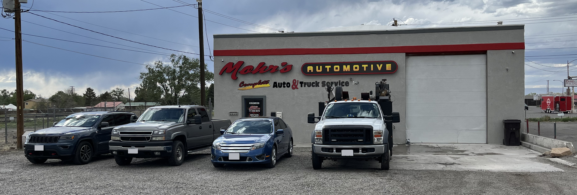 An automotive shop with four vehicles parked in front. Gray building with a red stripe, cloudy sky. | Mohr's Automotive