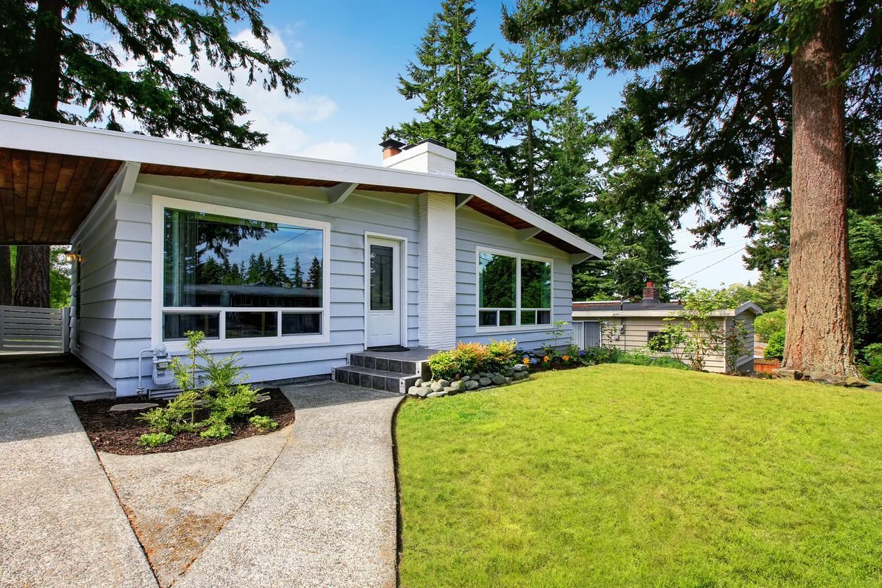 Light blue house with large windows, pathway, and green lawn. Tall trees in background.