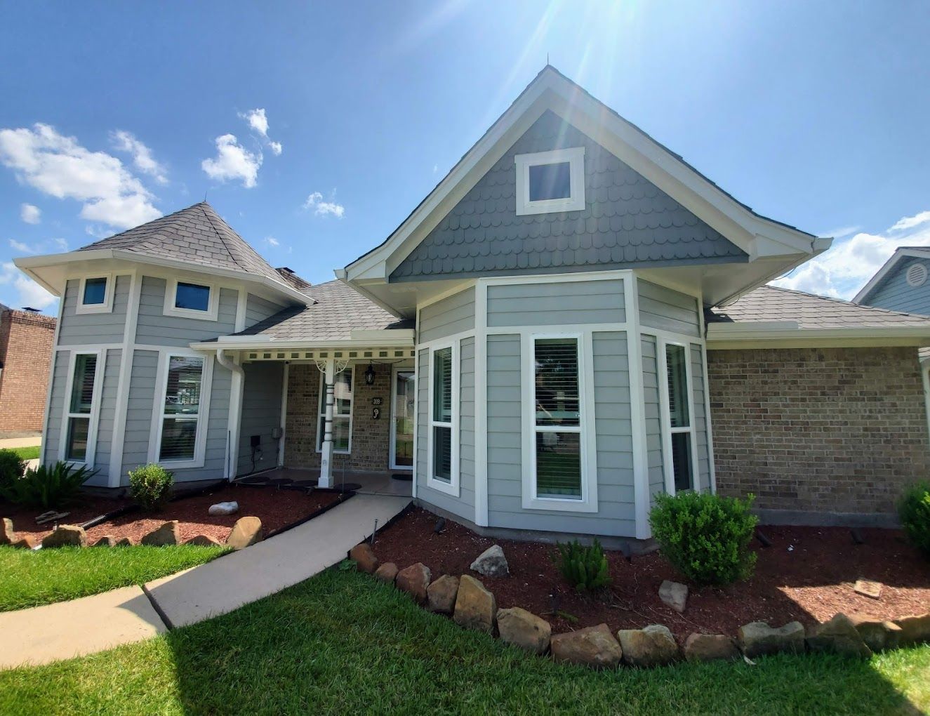 Light blue house with brown roof, windows, and manicured landscaping under a sunny sky.