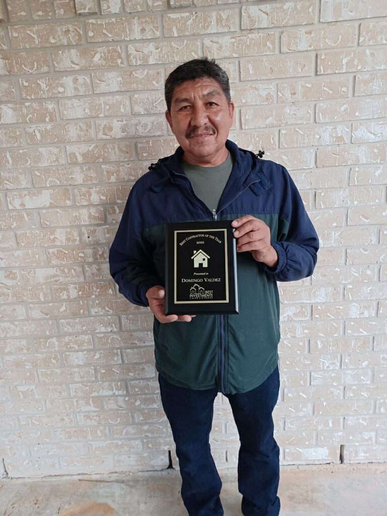 Man holding a plaque in front of a brick wall. He is wearing a jacket and jeans.