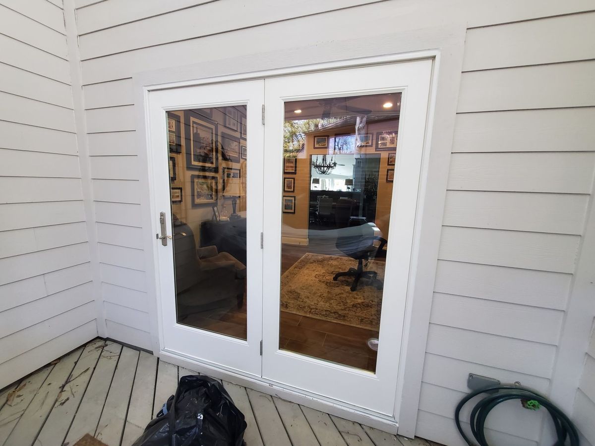 White French doors reflect a room with framed pictures and a rug. The doors are set in white siding, on a wooden deck.