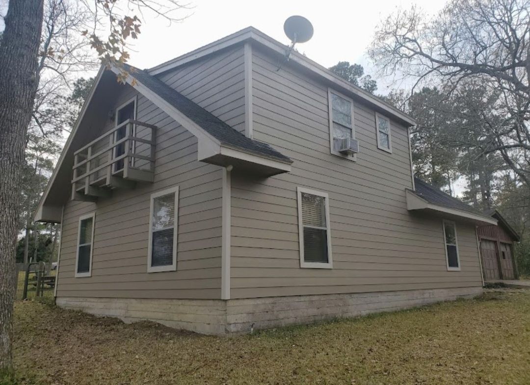 Tan two-story house with a balcony and satellite dish, surrounded by trees and grass.