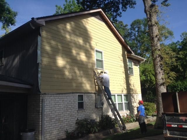Two workers on a ladder installing siding on a two-story yellow house with a brick base.