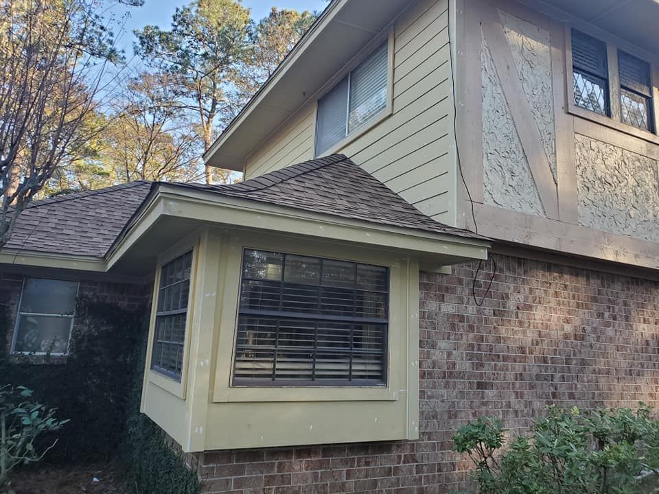 Brick and tan house exterior with bay window and roof. Trees in background.