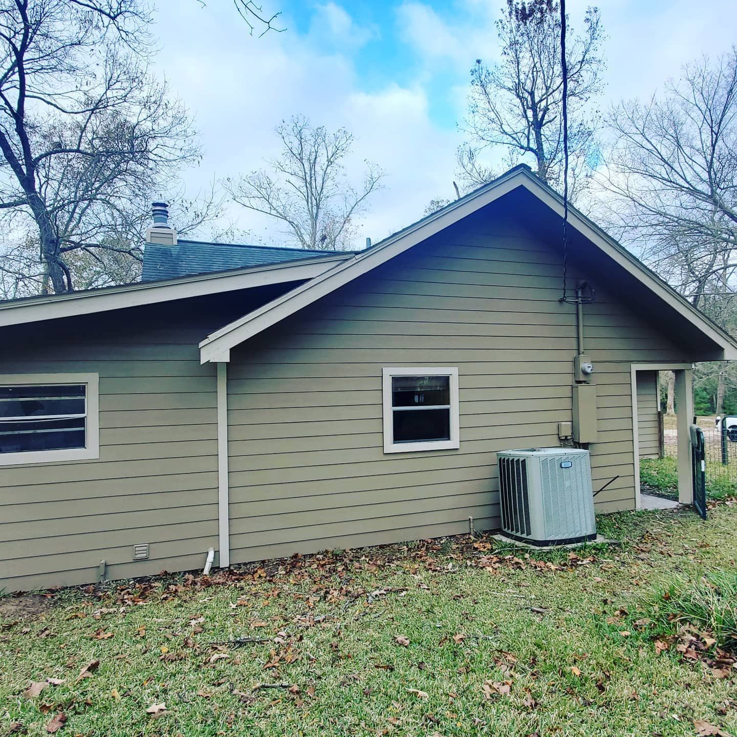 Tan house exterior with air conditioner, windows, and bare trees under a cloudy sky.
