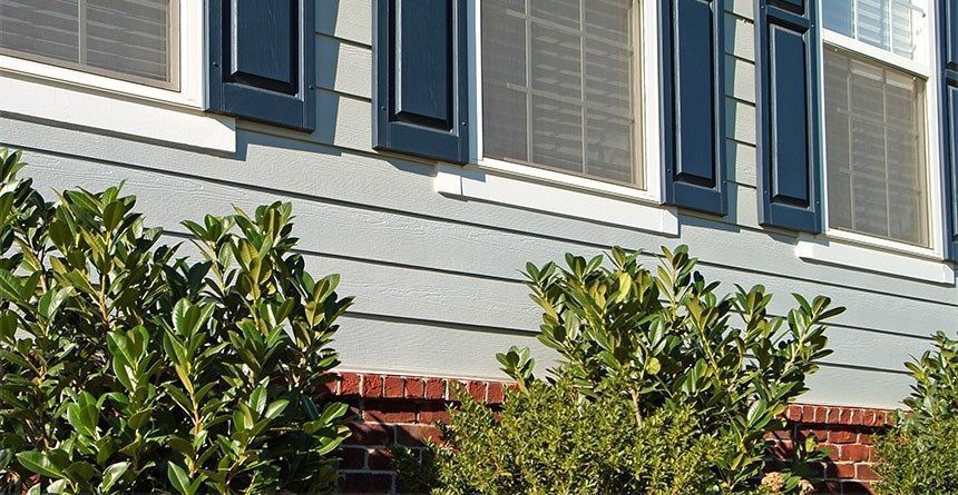 Gray house exterior with blue shutters, windows, and brick base; bushes in foreground.