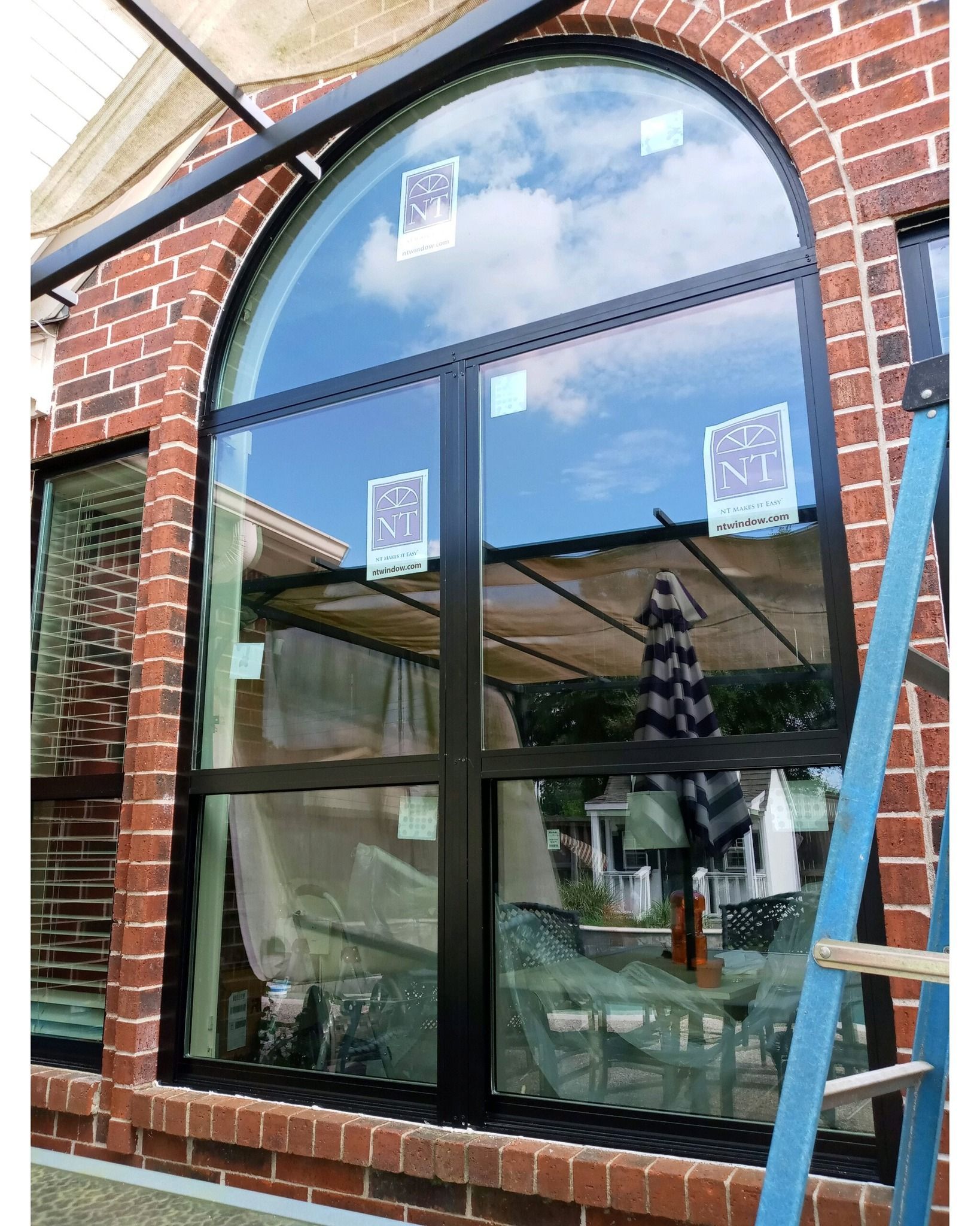 Large arched window in brick building, black frame, blue ladder, partially clouded sky reflection.