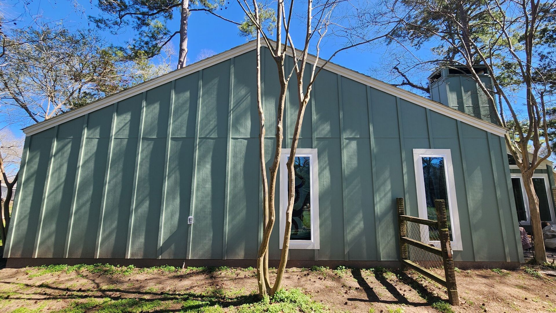 Green-sided building with two tall windows and a chimney, set amidst trees under a blue sky.