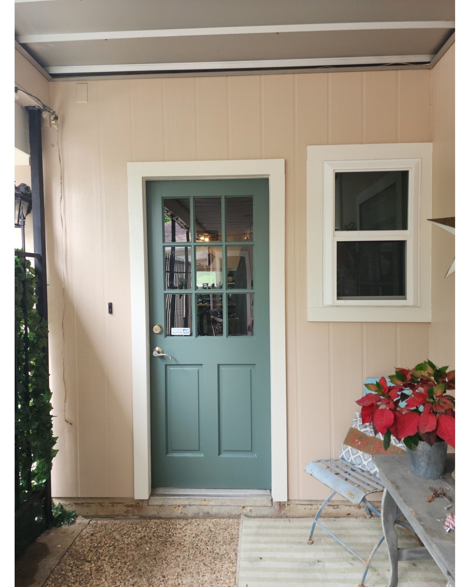 Green door with glass panes and a small window in a peach-colored exterior with a small bench and potted plants.
