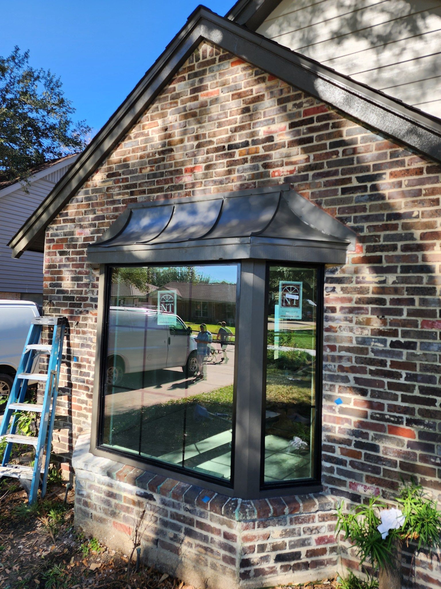 Bay window with dark trim and roof installed on a brick building with a ladder nearby.