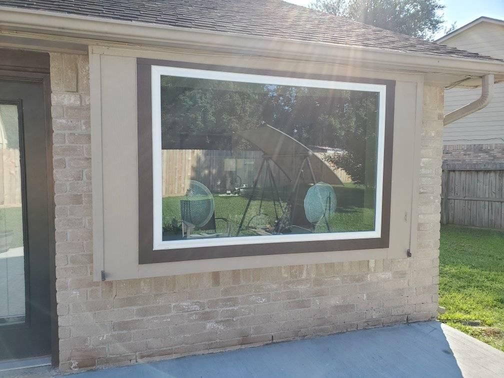A large rectangular window on a brick building with a brown frame, reflecting a sunny backyard.