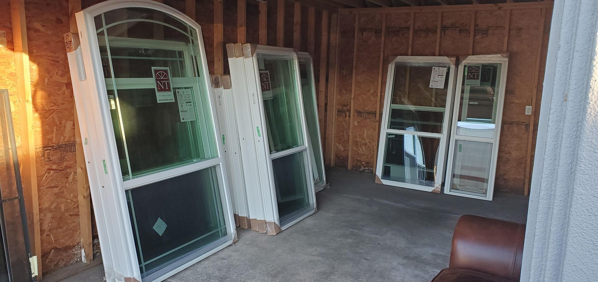 Several white-framed windows lean against a wood-paneled wall, in a storage area. A brown chair sits nearby.