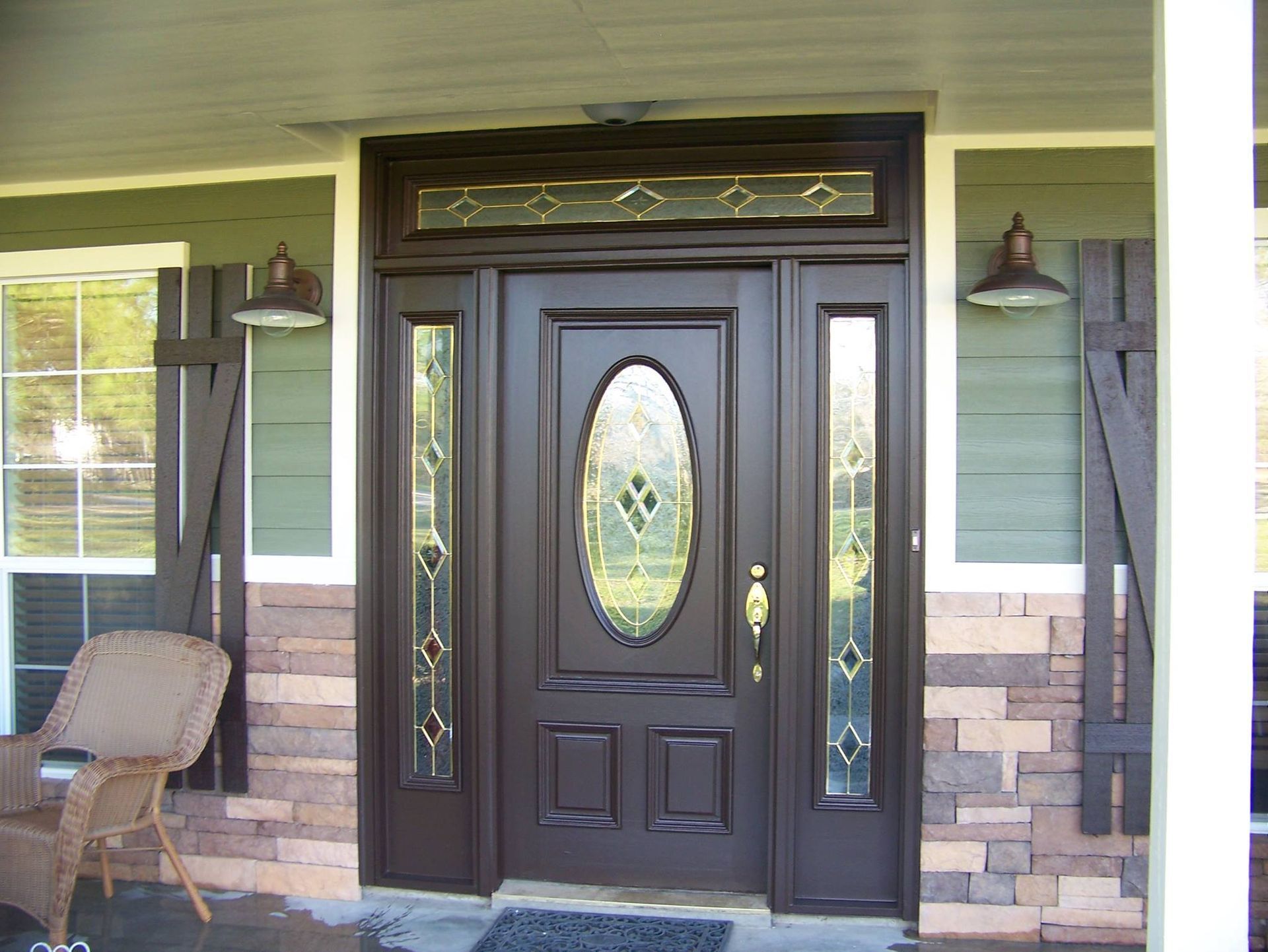 Dark brown front door with stained glass, sidelights, and transom; flanked by sconces and shutters.