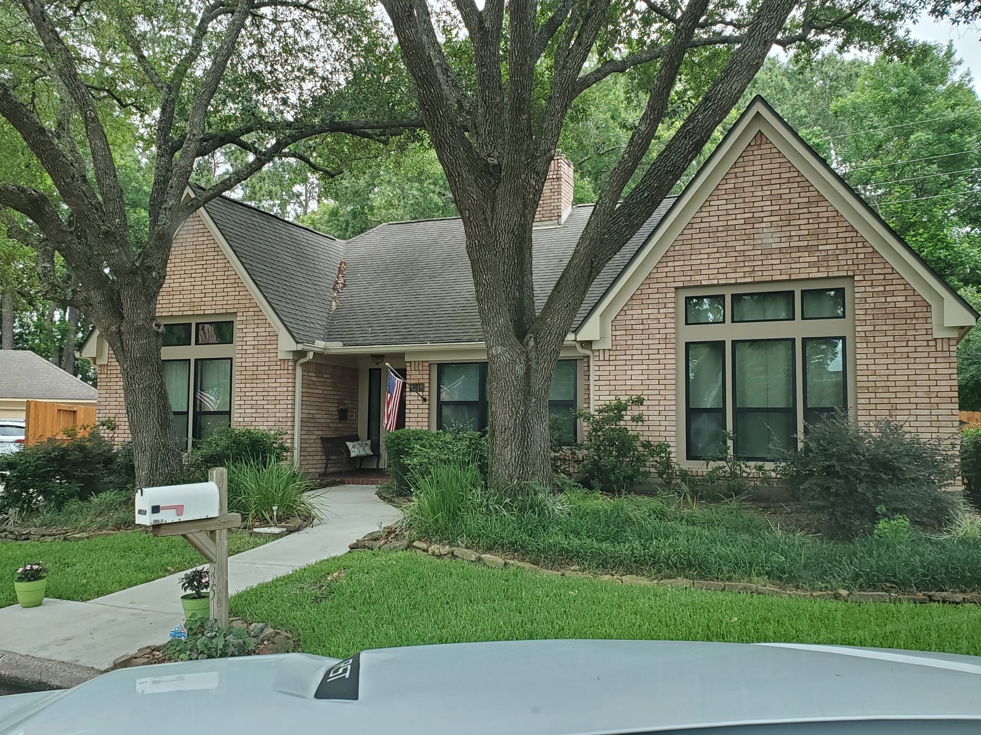 Brick house with a dark roof and large trees in front; mailbox and sidewalk visible.