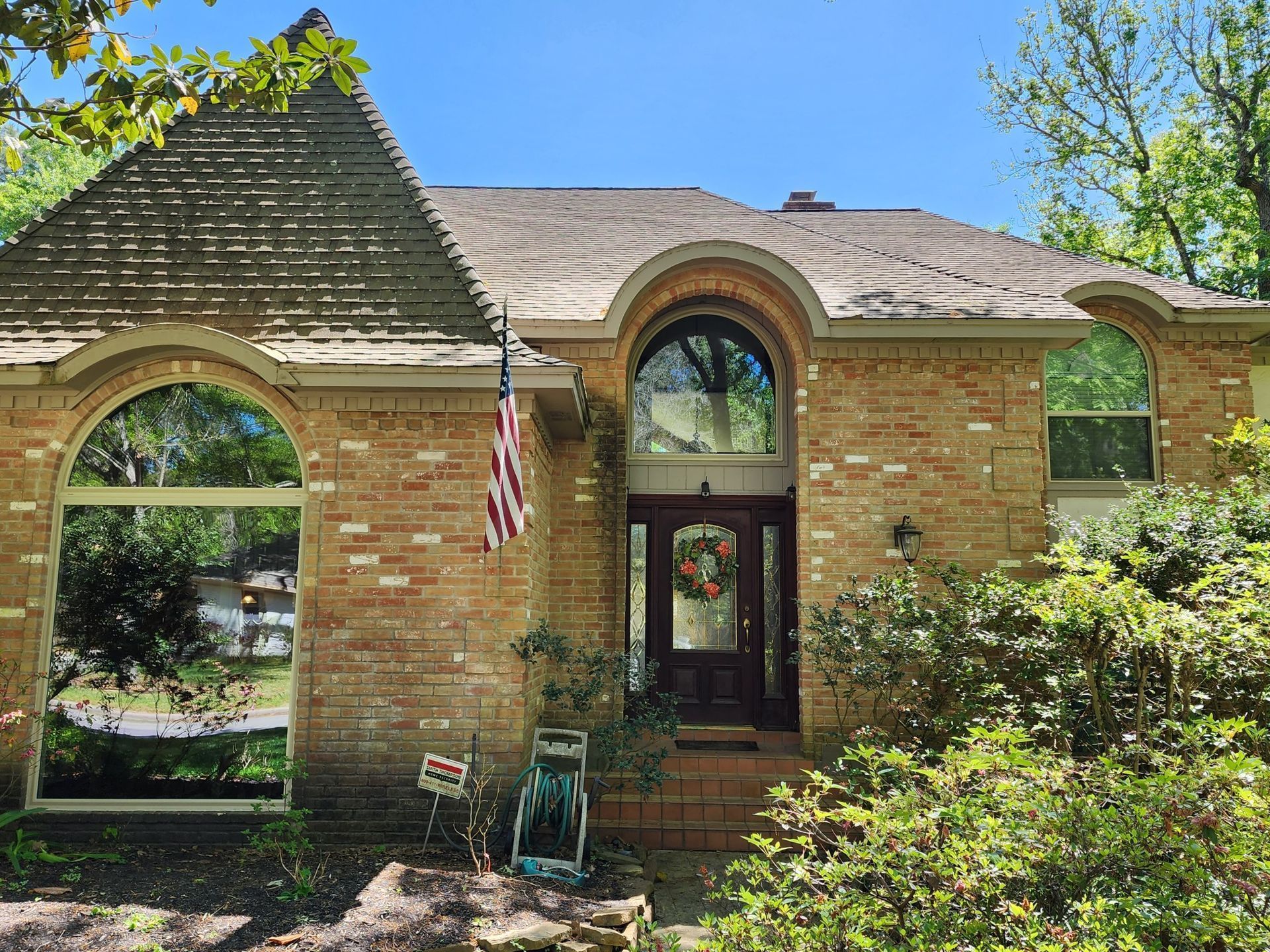 Brick house with arched windows, wooden door, American flag, and surrounding greenery under a blue sky.