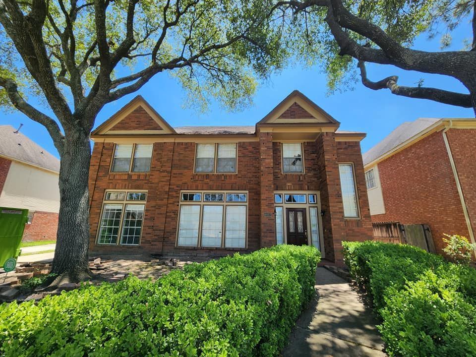 Brick house with two trees and a well-kept yard. Blue sky above, bushes below.