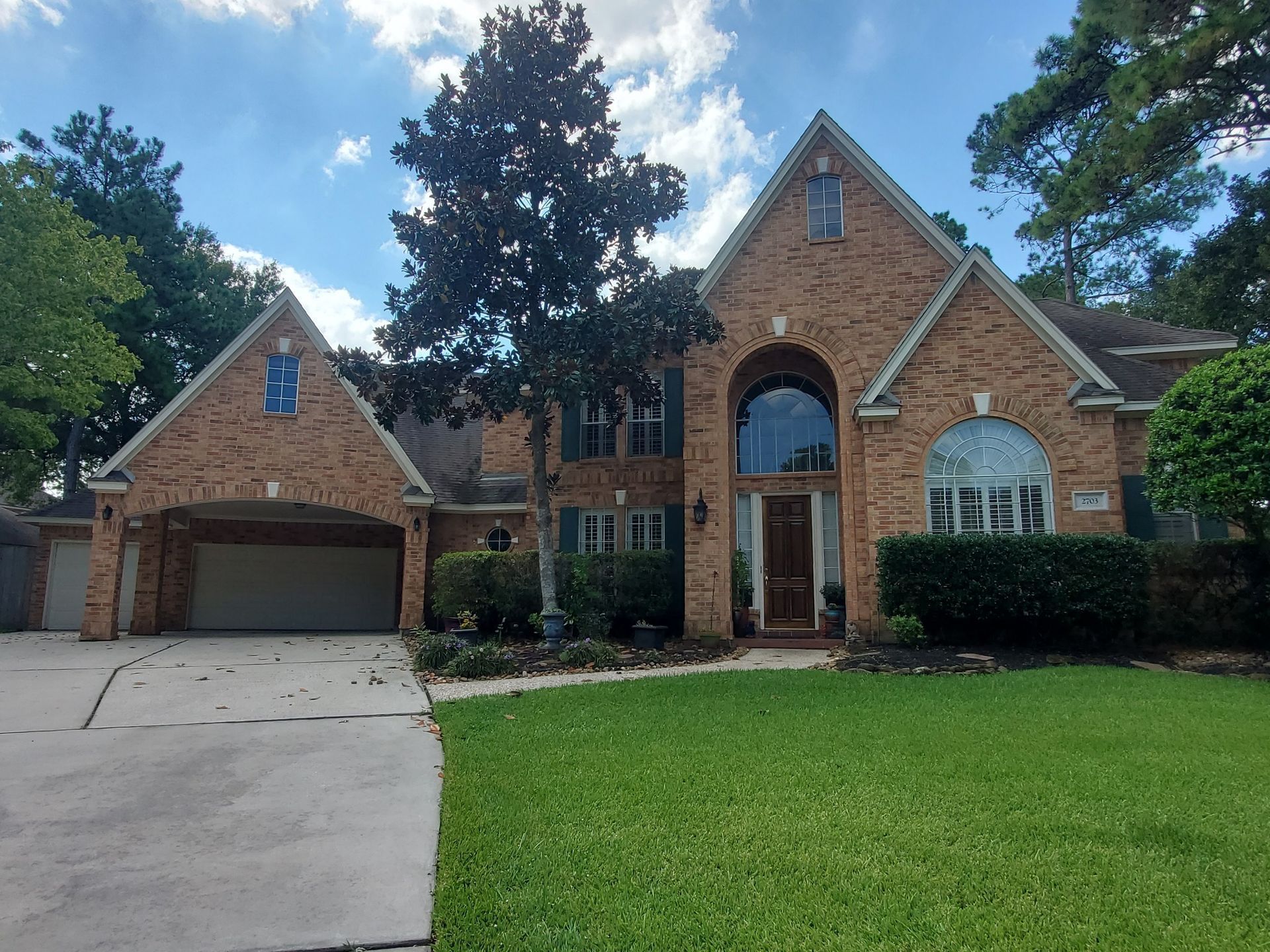 Two-story brick house with arched entrance and attached garage; green lawn, trees, blue sky.