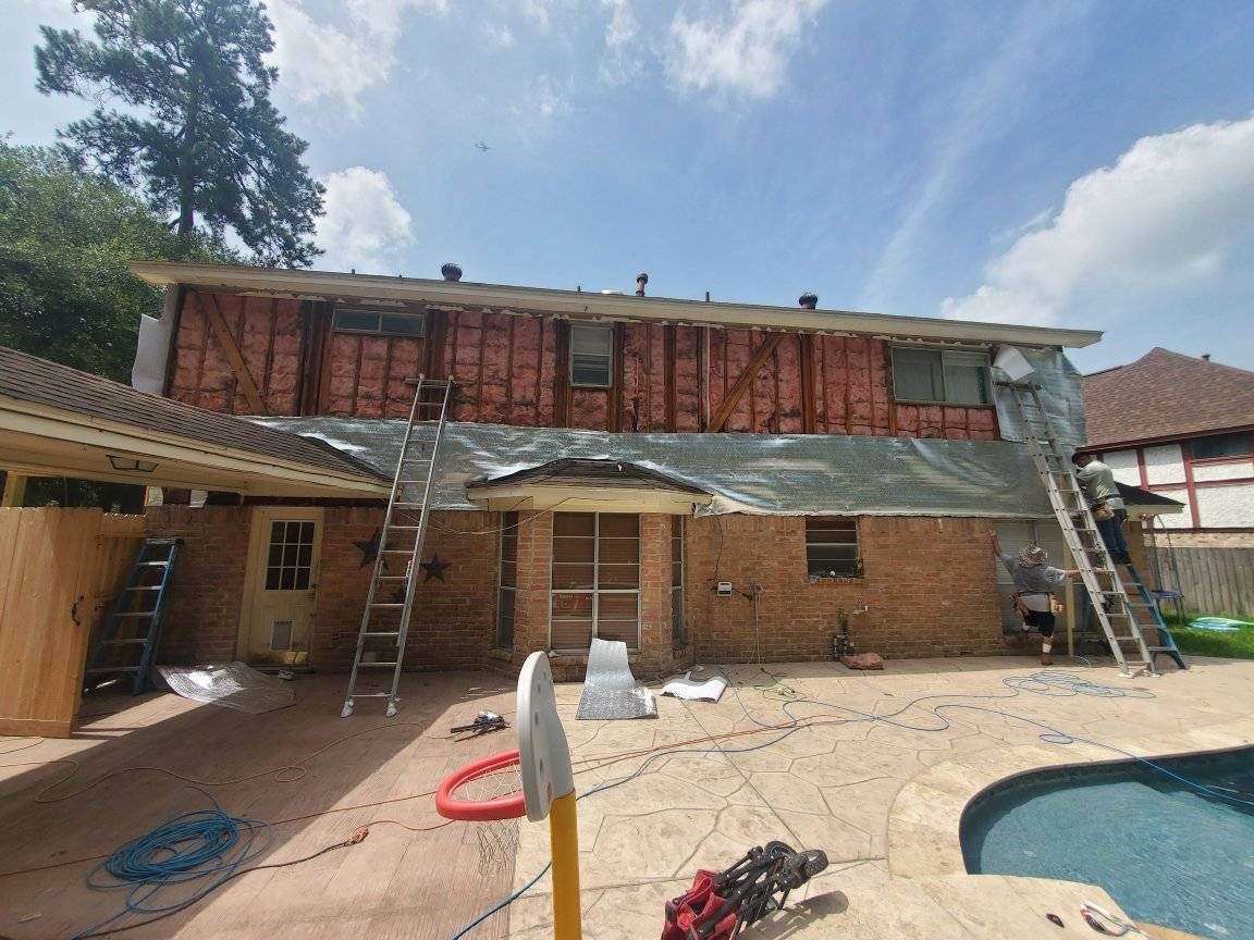 Workers renovating a two-story brick house with exposed wall framing. Ladders, tarp, and insulation are visible on a sunny day.