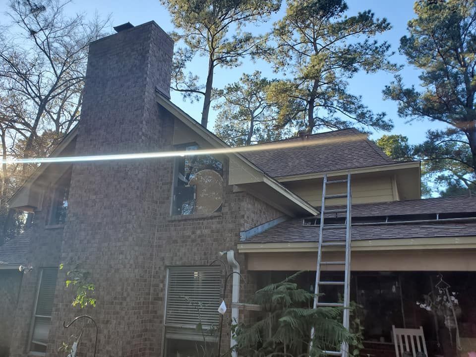 Brick house with chimney, ladder on roof, surrounded by trees and sunny sky.