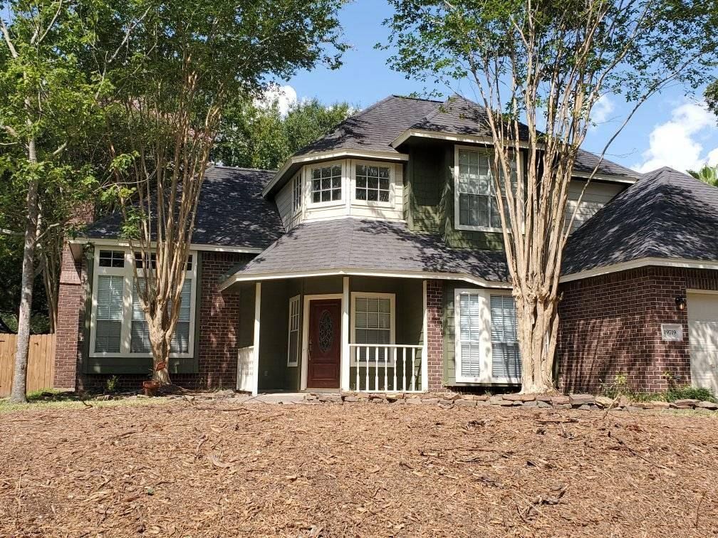 Two-story house with green siding, brick accents, and dark roof. Trees in front. Sunny day.