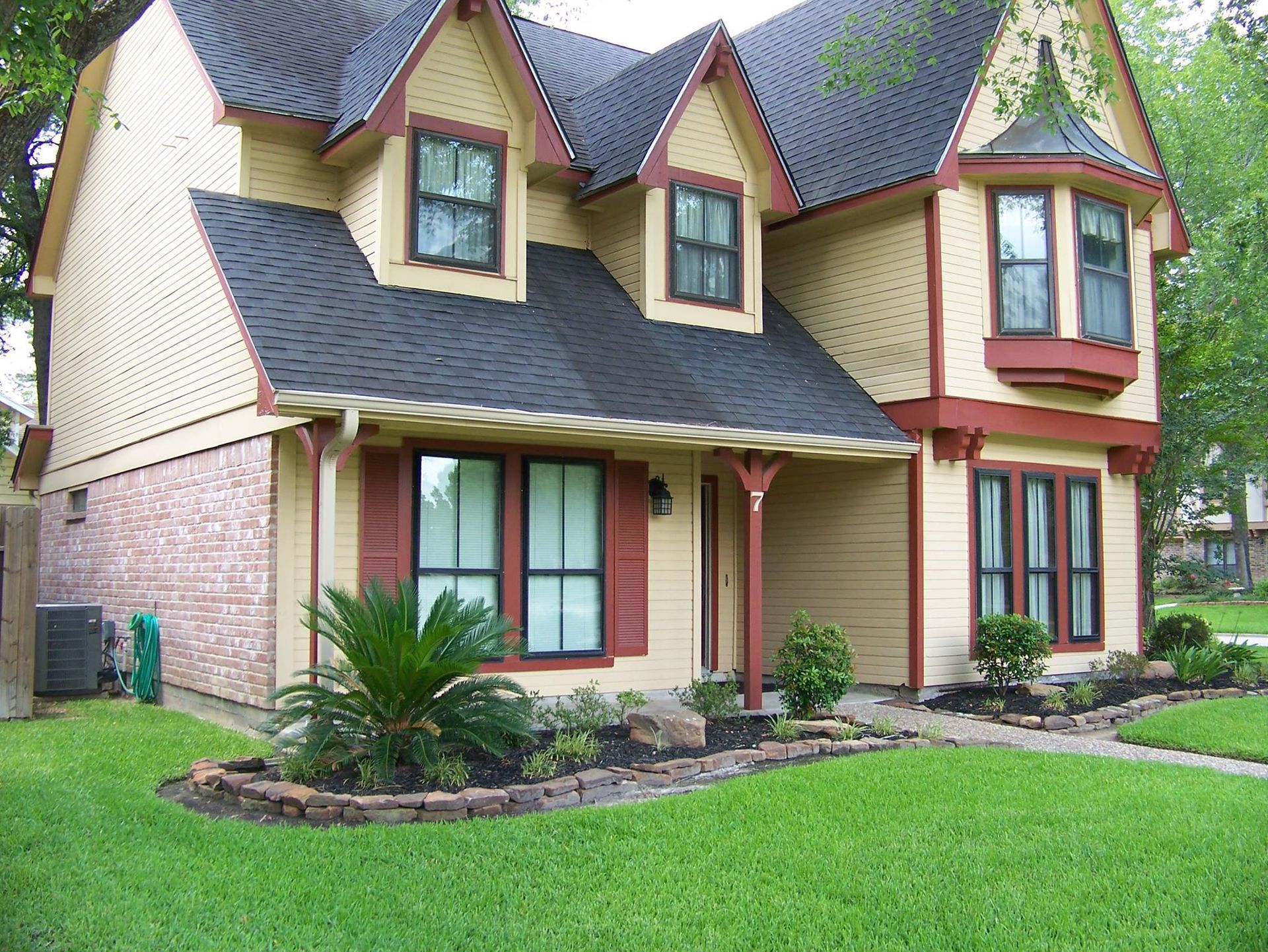 Two-story house with tan siding, burgundy trim, and a black roof. Lush green lawn and garden in front.