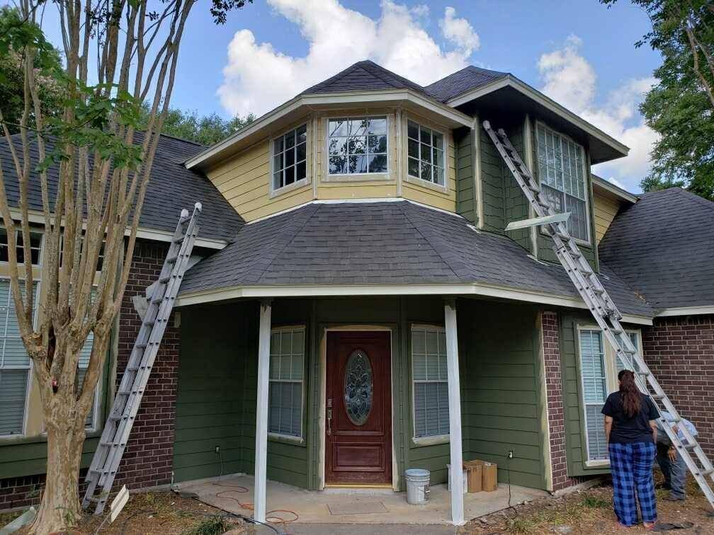 House exterior being painted; ladders propped up against the building, person standing nearby, blue sky.