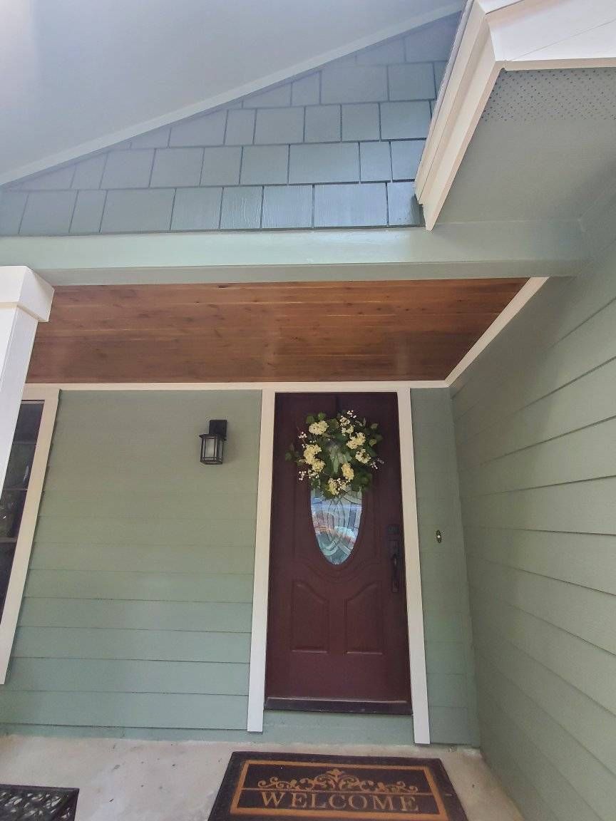 Green-painted house entryway with a burgundy door and a sunflower wreath, beneath a wood-paneled ceiling.