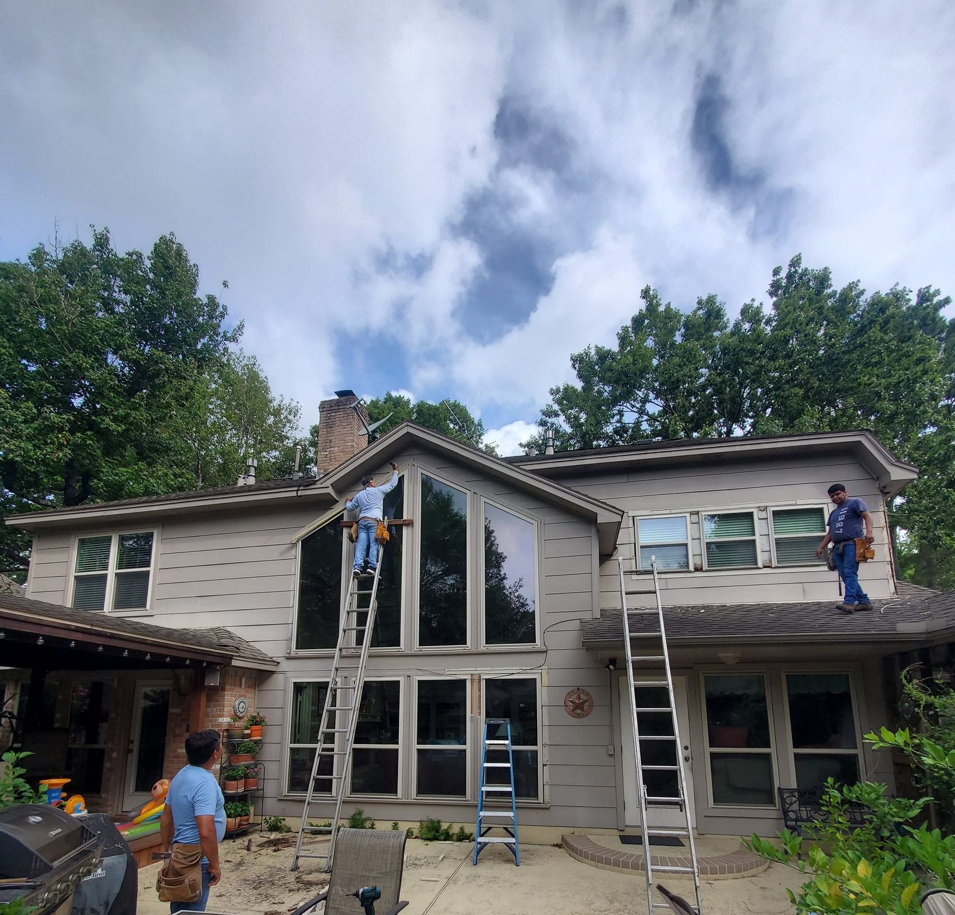Workers on ladders replacing siding on a two-story house with tall windows under a cloudy sky.