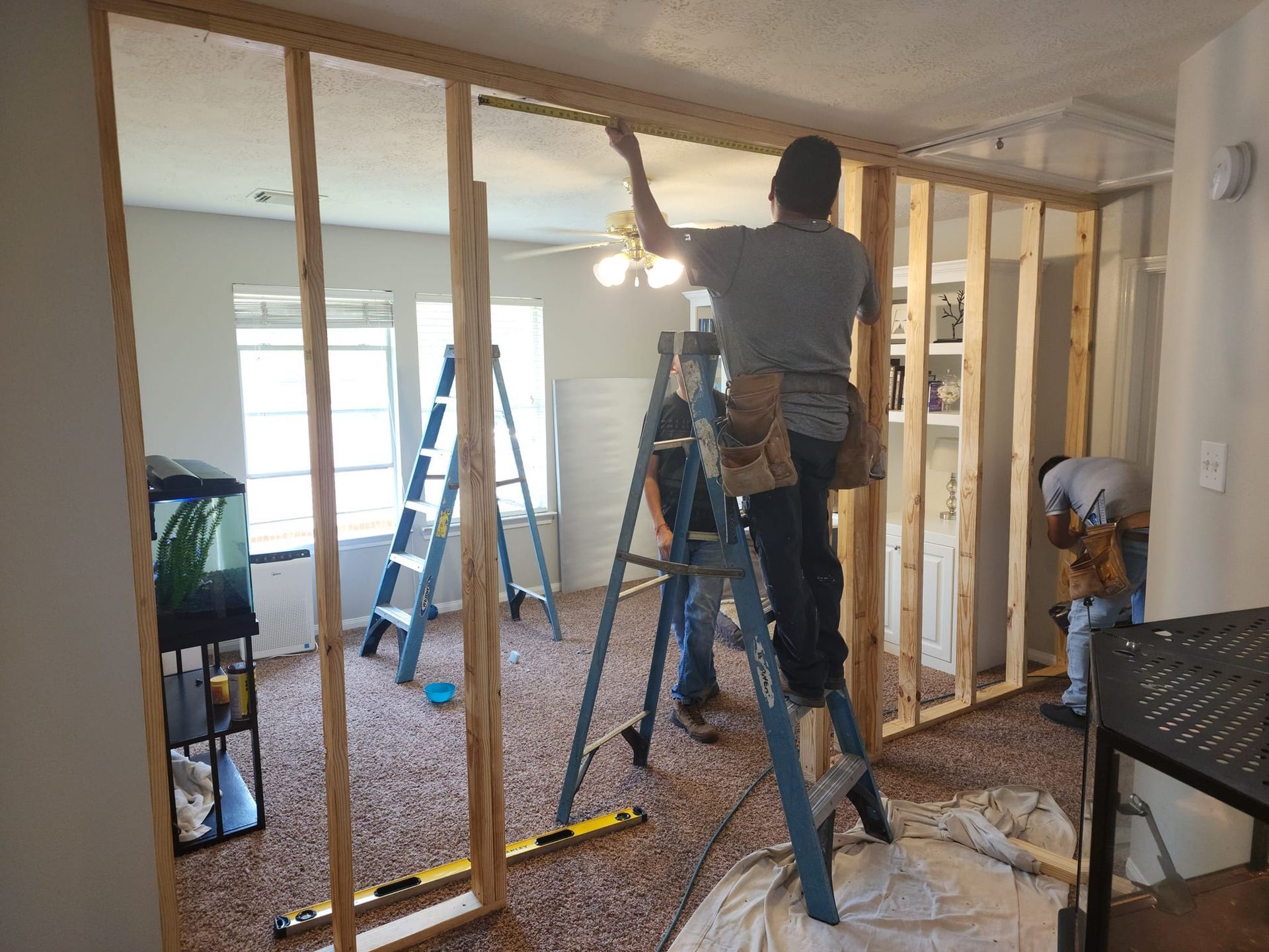 Men building a wooden wall inside a home; one on a ladder, others assisting.
