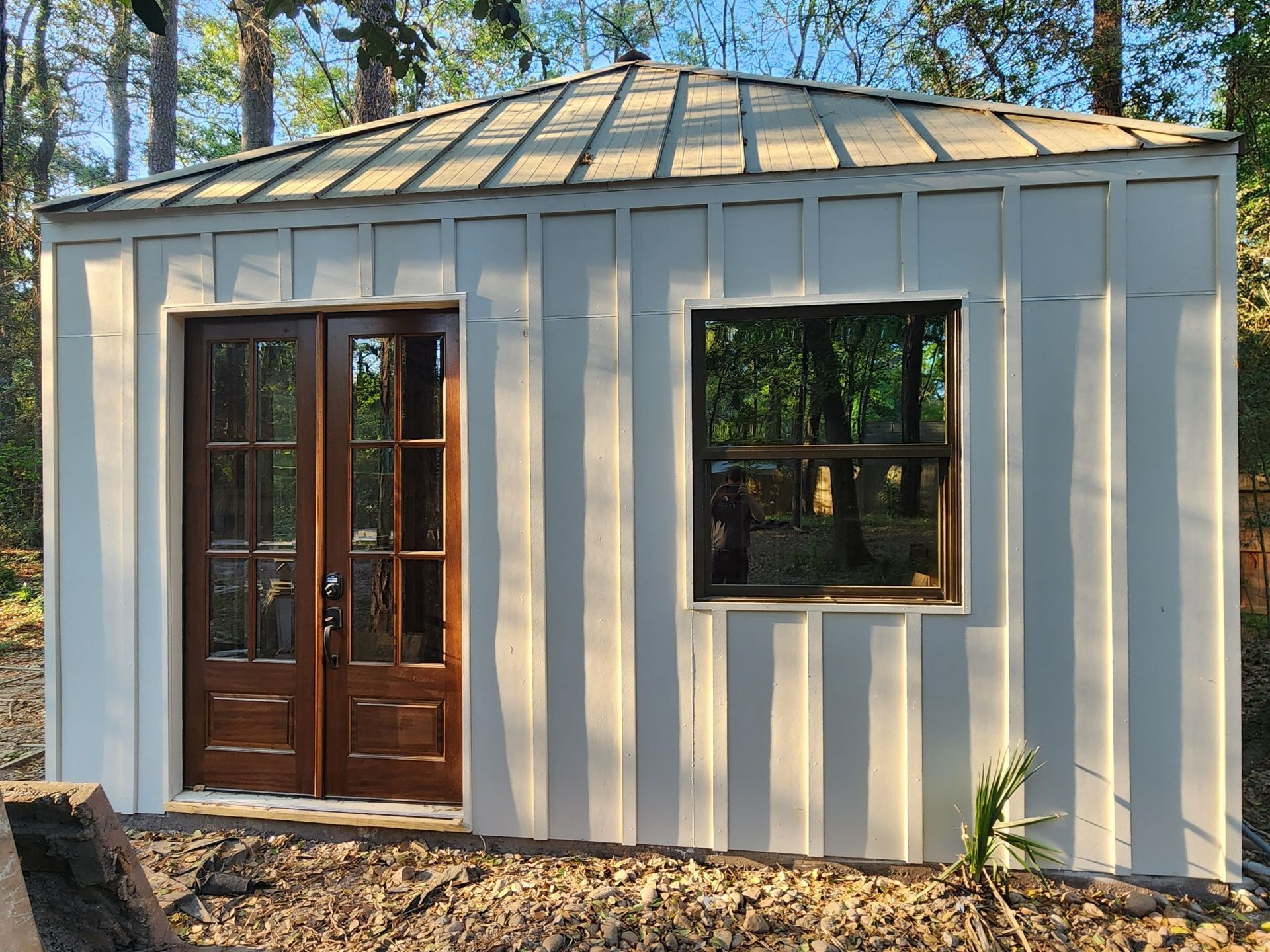 White shed with brown double doors and window, surrounded by trees.
