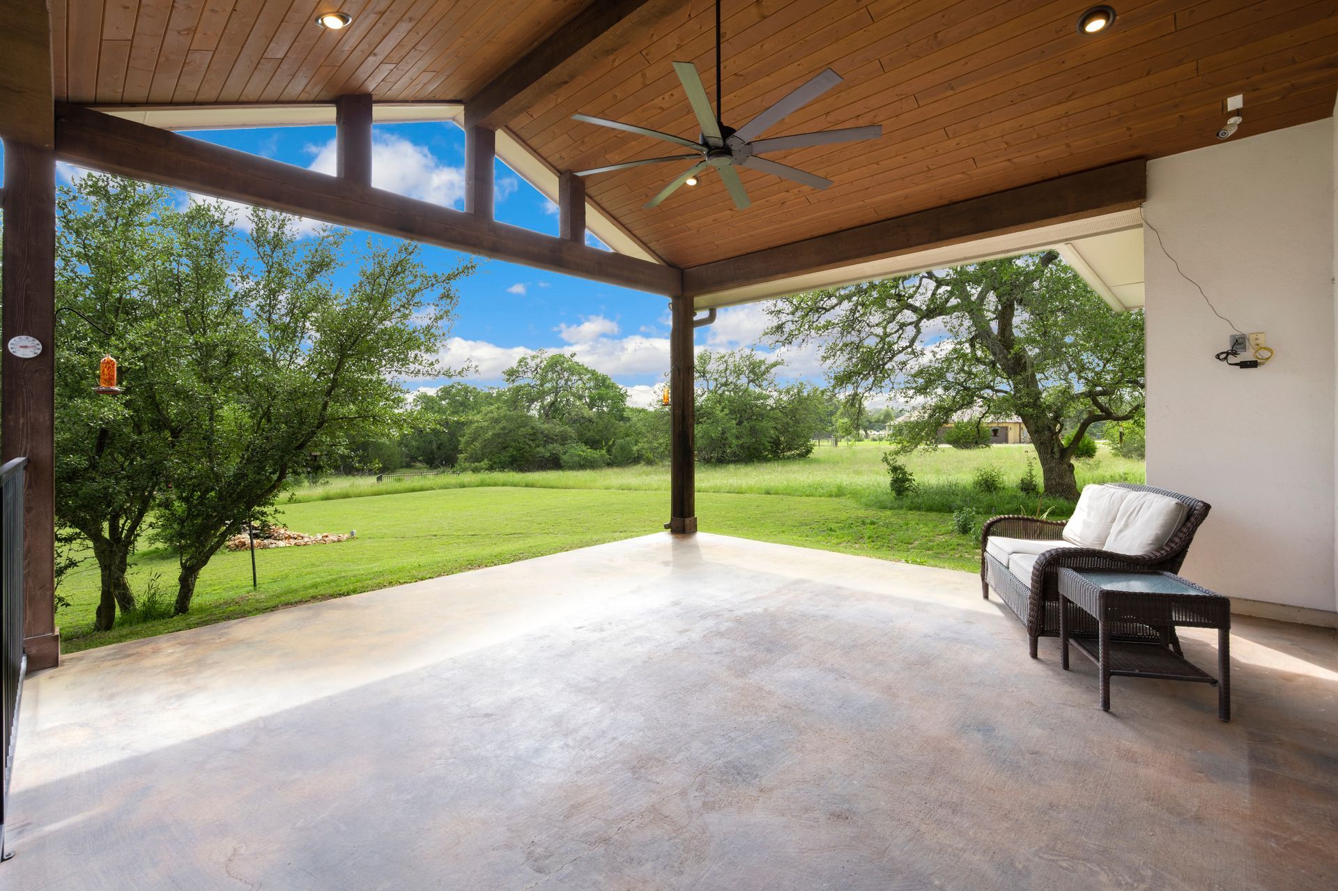 Covered patio with a view of a green field and trees under a blue sky. Furnished with a chair and side table.