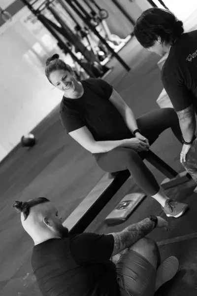People in a gym; two looking at an item while another sits on a bench, smiling. Black and white.