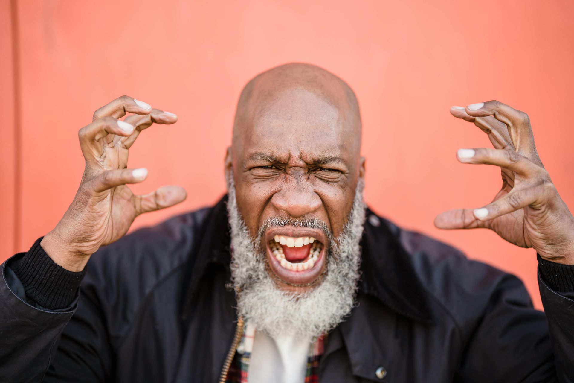 An angry Black man with a shaved head and gray beard yells, hands raised, against an orange background.