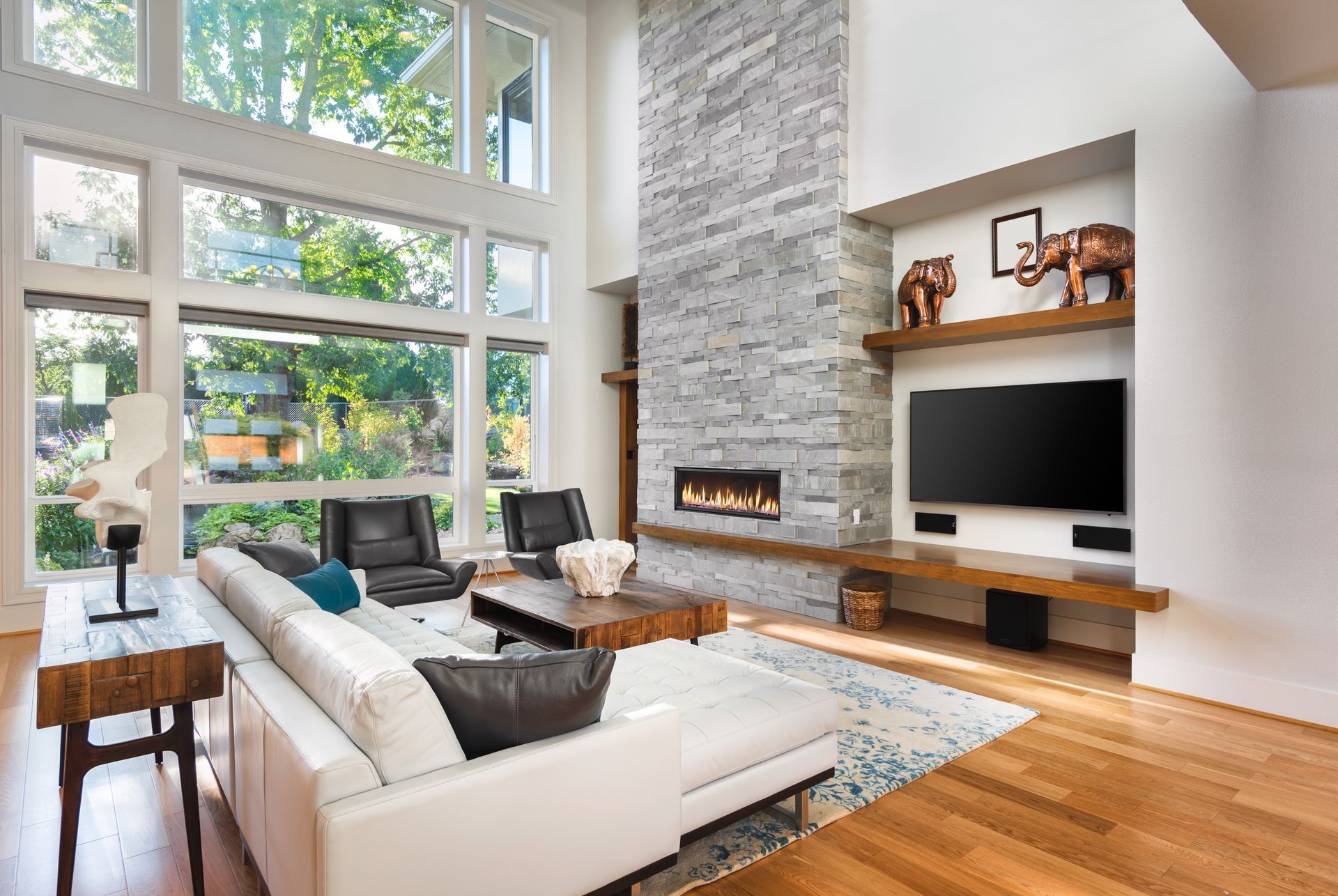 Bright modern living room with floor-to-ceiling windows, stone fireplace, white sofas, and wood floors