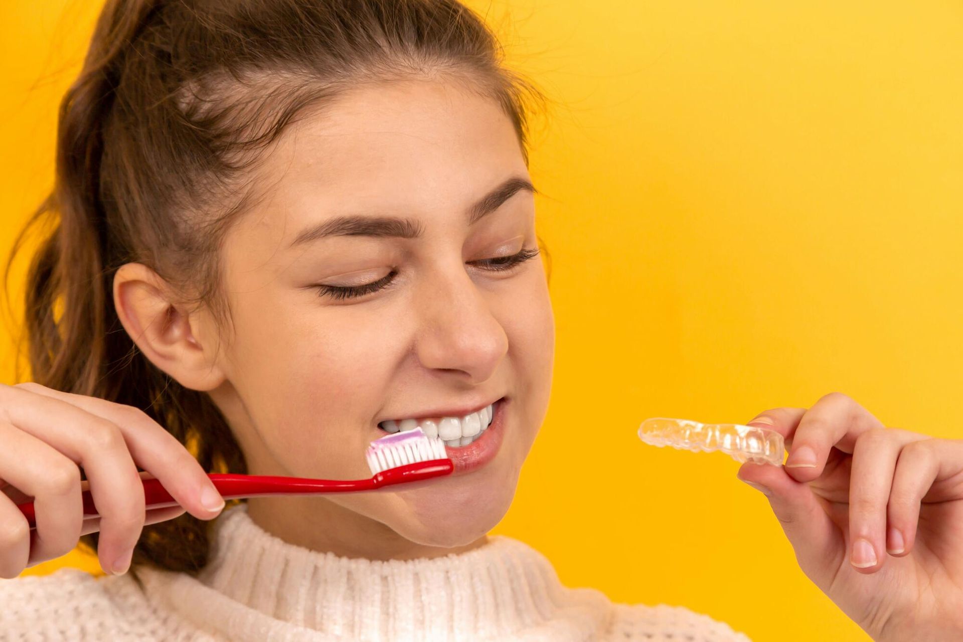 Woman brushing teeth with Invisalign aligner in hand, yellow background.