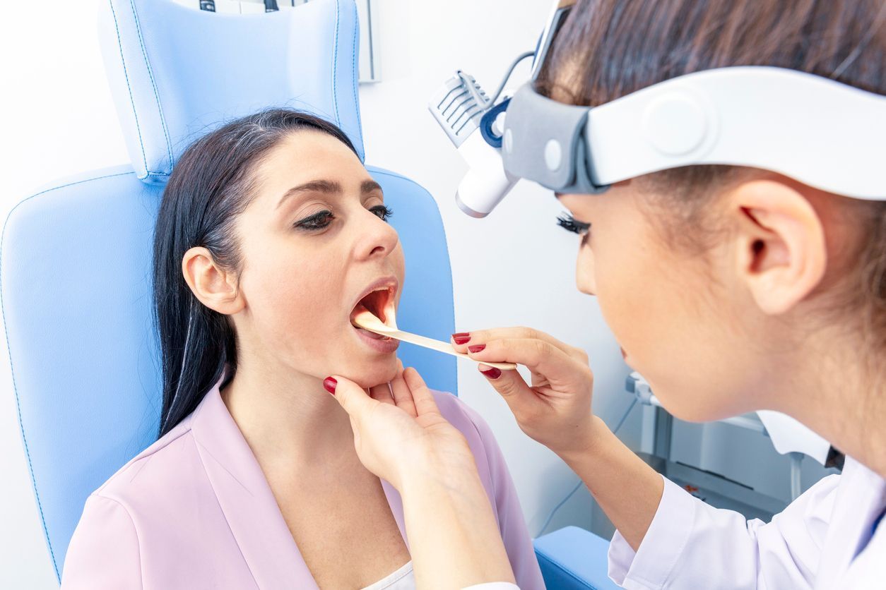 A healthcare professional using a tongue depressor and headlamp to examine a patient's throat in a clinical setting.