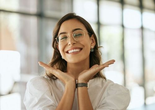 A woman wearing glasses is smiling with her hands on her face.