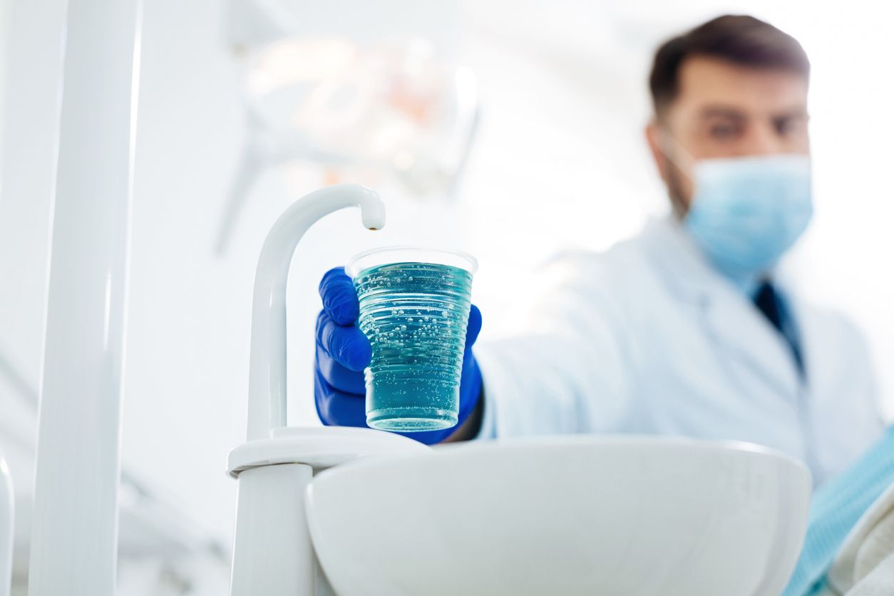 Dentist in blue gloves holds a cup of blue liquid over a sink in a dental office.
