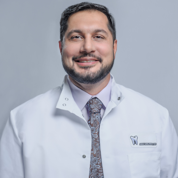 Smiling man in a white lab coat with a tie. Likely a dentist.