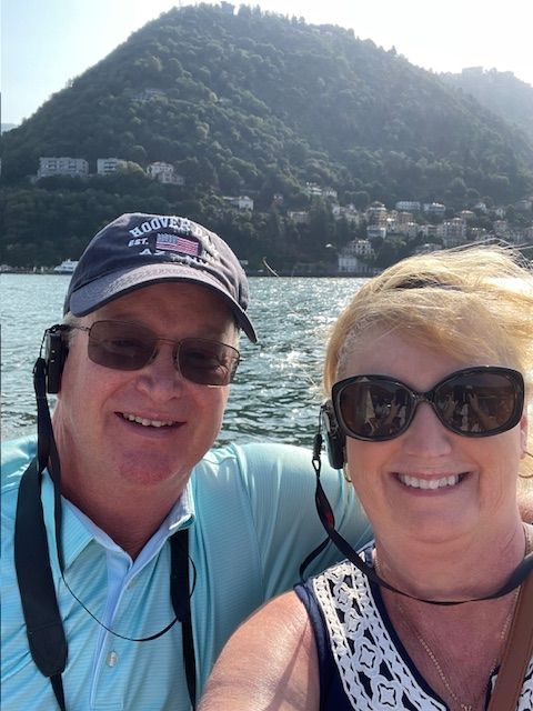 A smiling couple in sunglasses takes a selfie on a boat with a mountainous, tree-covered shoreline in the background.