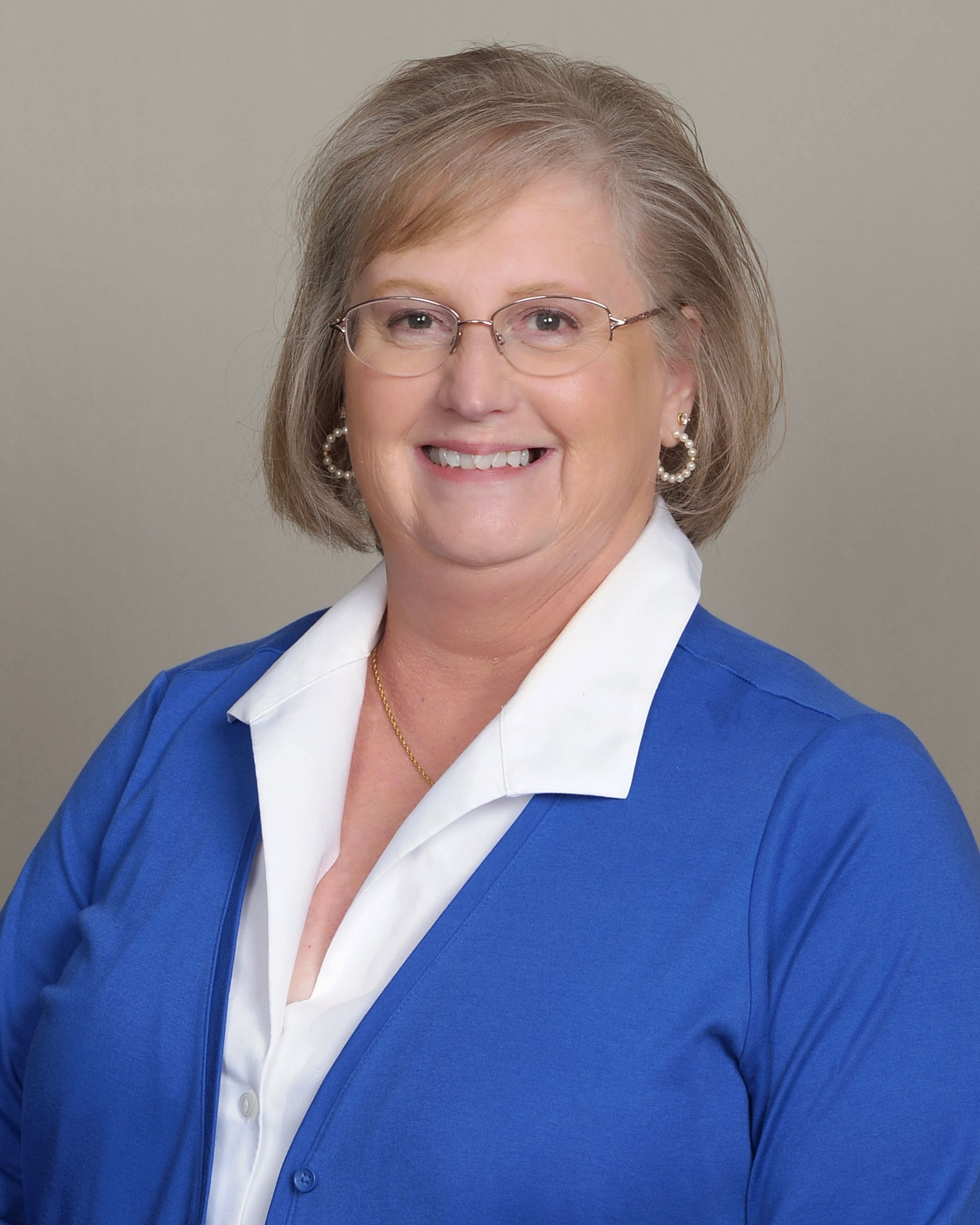 A smiling person with shoulder-length blonde hair and glasses wearing a blue cardigan over a white collared shirt.