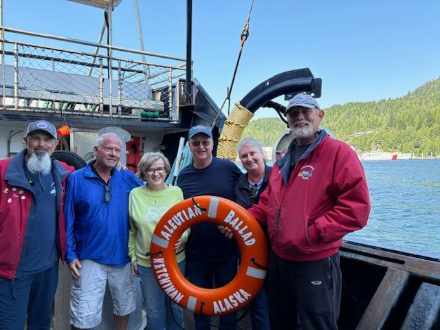 Six people stand on a boat deck in Ketchikan, Alaska, holding a life ring labeled Aleutian Ballad.