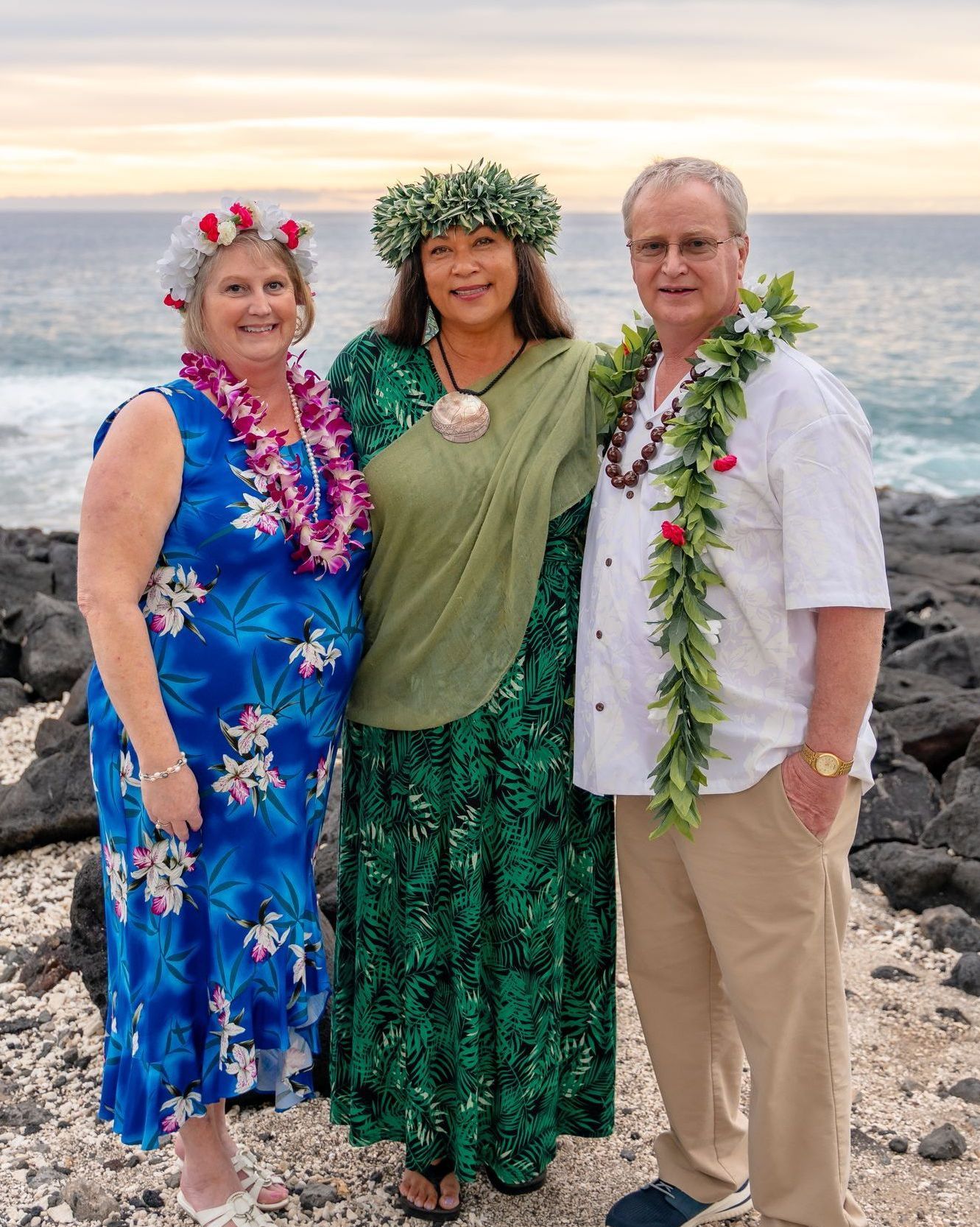 Three people in leis stand on a rocky beach at sunset, with two wearing patterned dresses and one in a white shirt.
